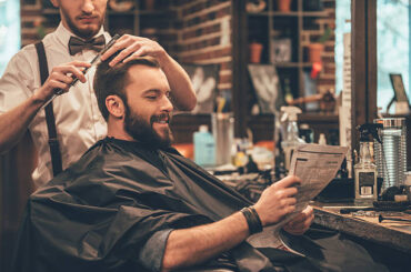 Cheerful young bearded man getting haircut by hairdresser and reading newspaper while sitting in chair at barbershop